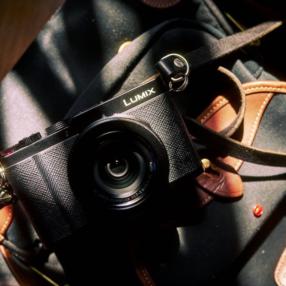 A black Lumix camera with a strap rests on a dark blue and brown bag in sunlight.