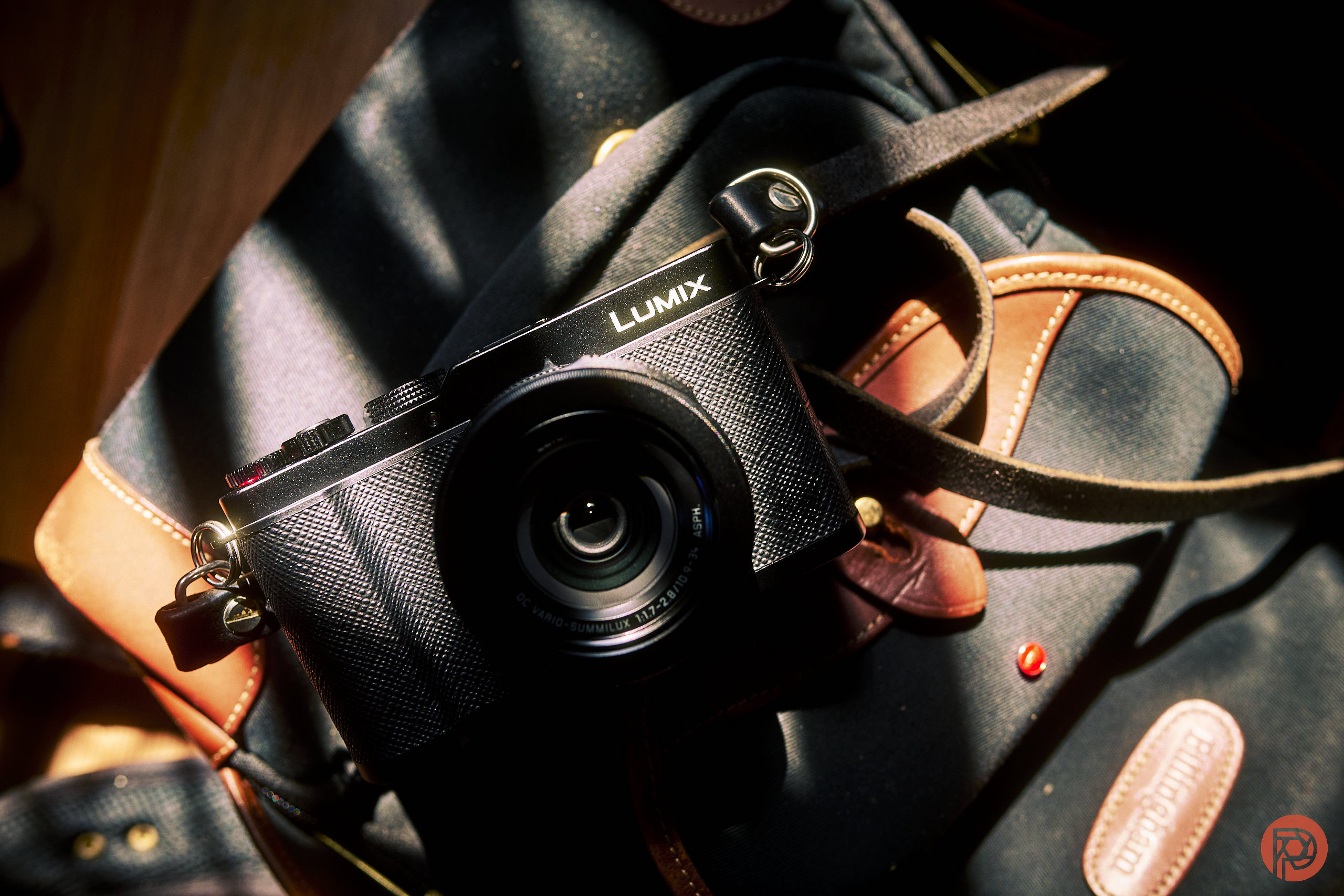 People are Searching for the Original Fuji X100. But Why? A black Lumix camera with a strap rests on a dark blue and brown bag in sunlight.