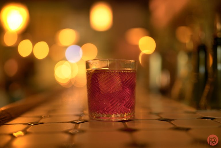 A cocktail glass with a pink drink sits on a tiled bar, with warm, blurry lights in the background.