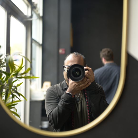 Person taking a photo in a mirror; another person and some plants are visible in the background.