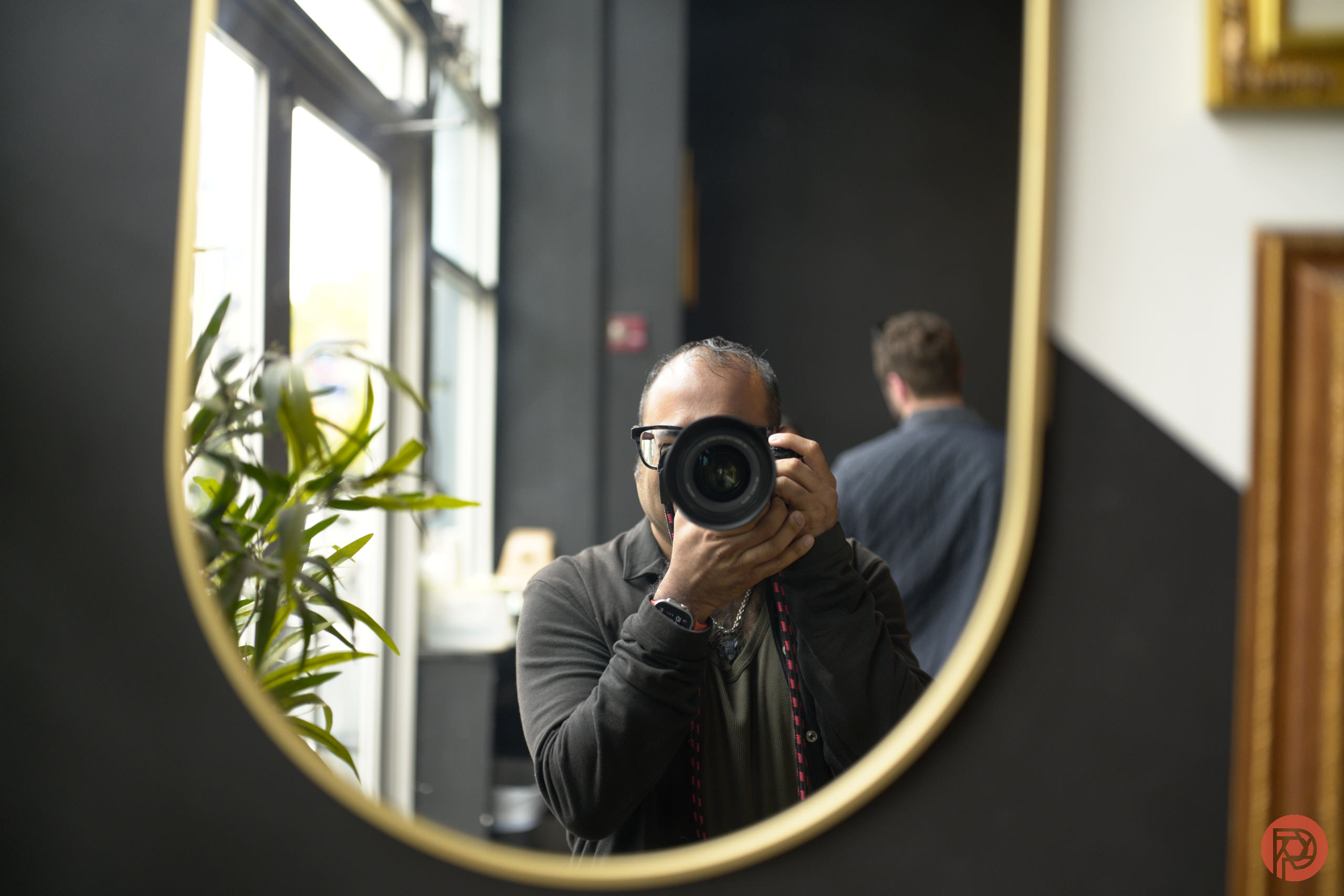 Person taking a photo in a mirror; another person and some plants are visible in the background.
