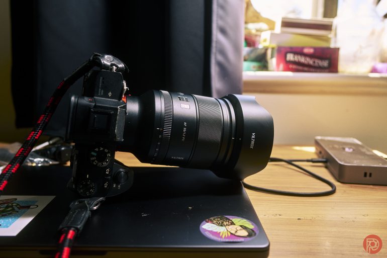A camera with a large lens rests on a closed laptop on a wooden desk near a window with books.