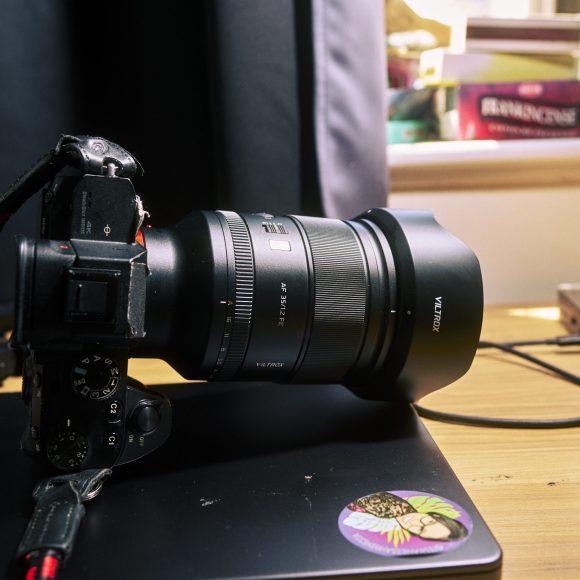 A camera with a large lens rests on a closed laptop on a wooden desk near a window.