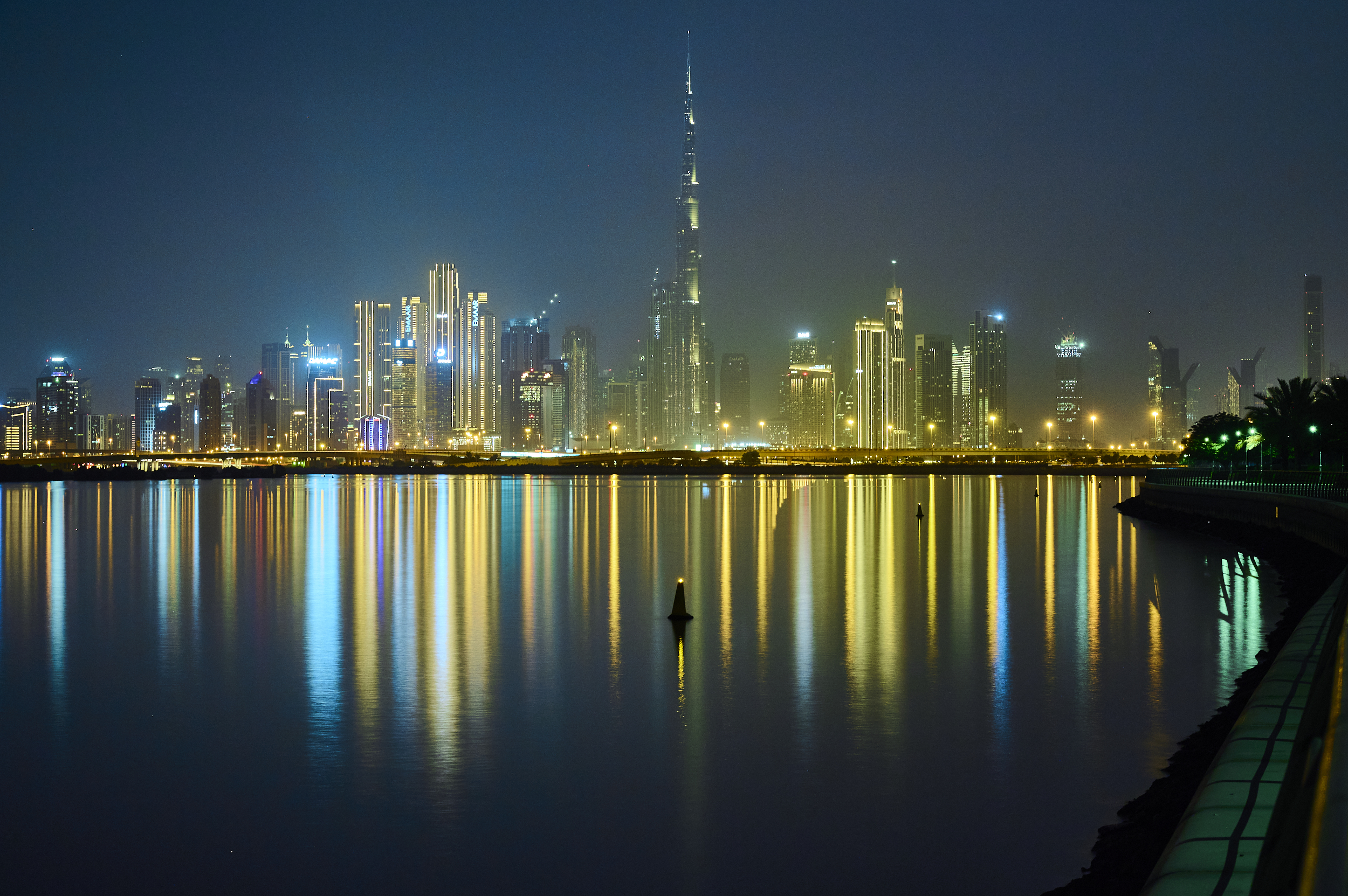 Dubai skyline at night, with colorful lights reflecting on calm water under a clear, dark sky.