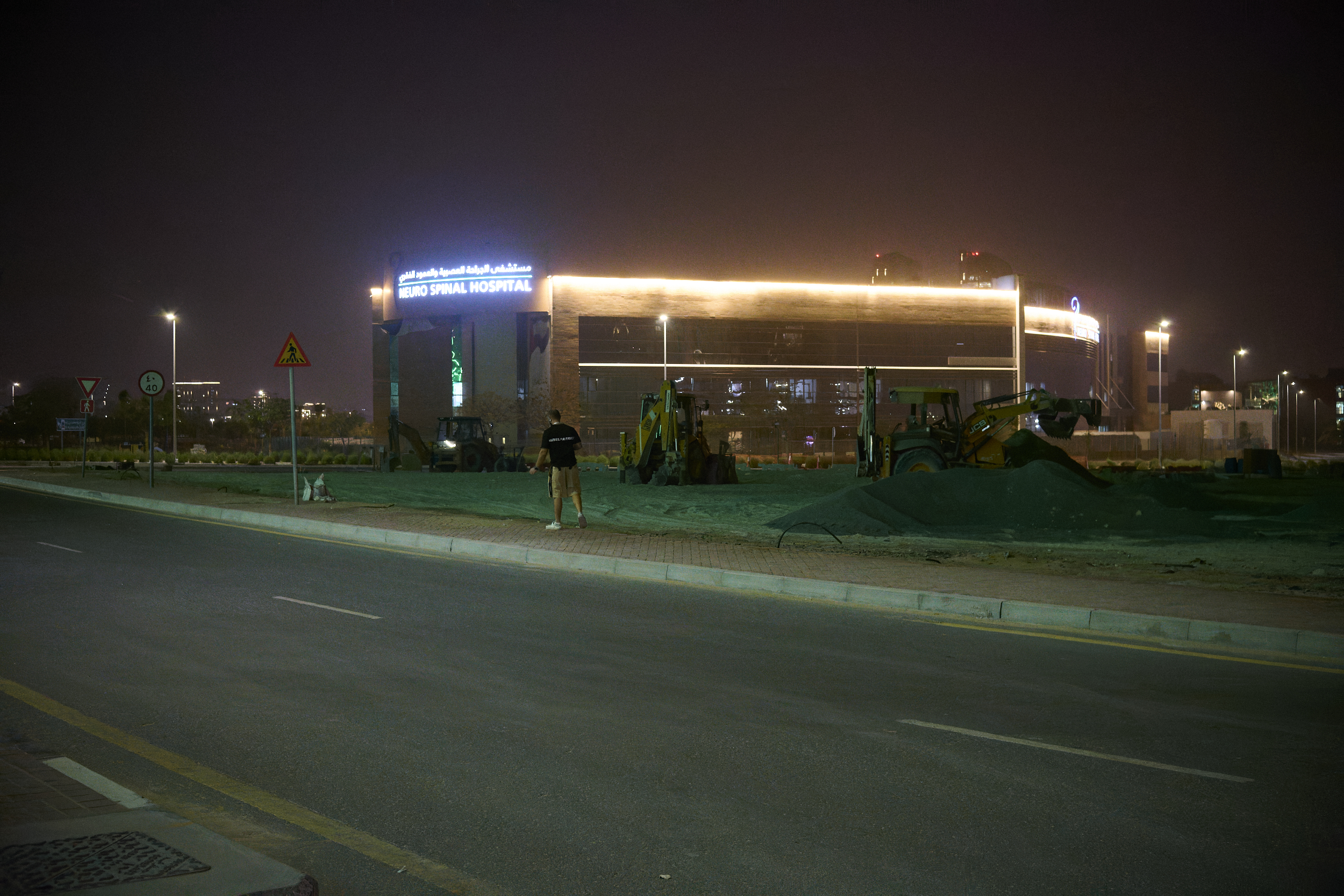 A person stands near construction equipment at night, with a brightly lit hospital in the background.