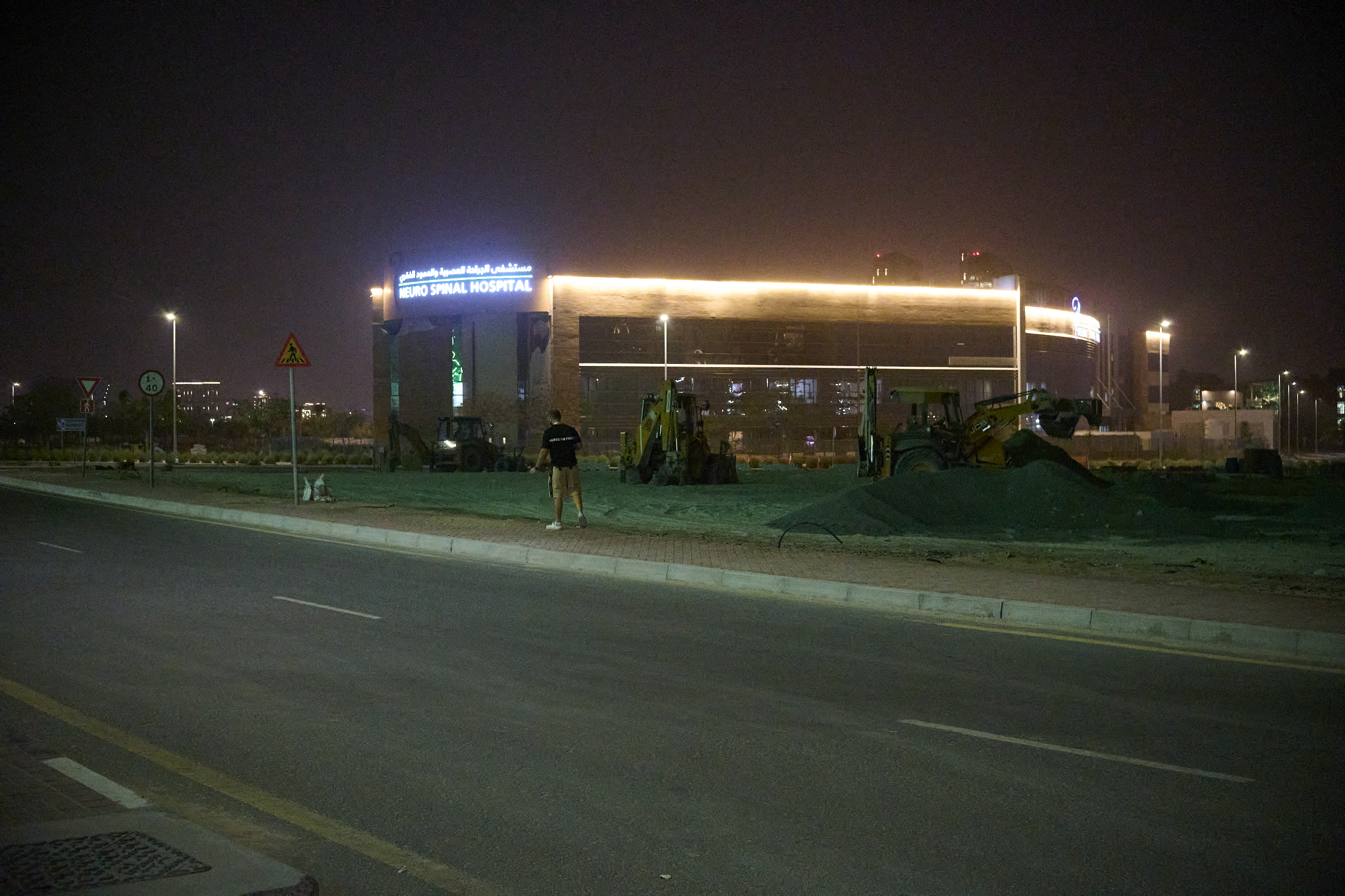 A person stands near construction equipment at night, with a lit hospital building in the background.
