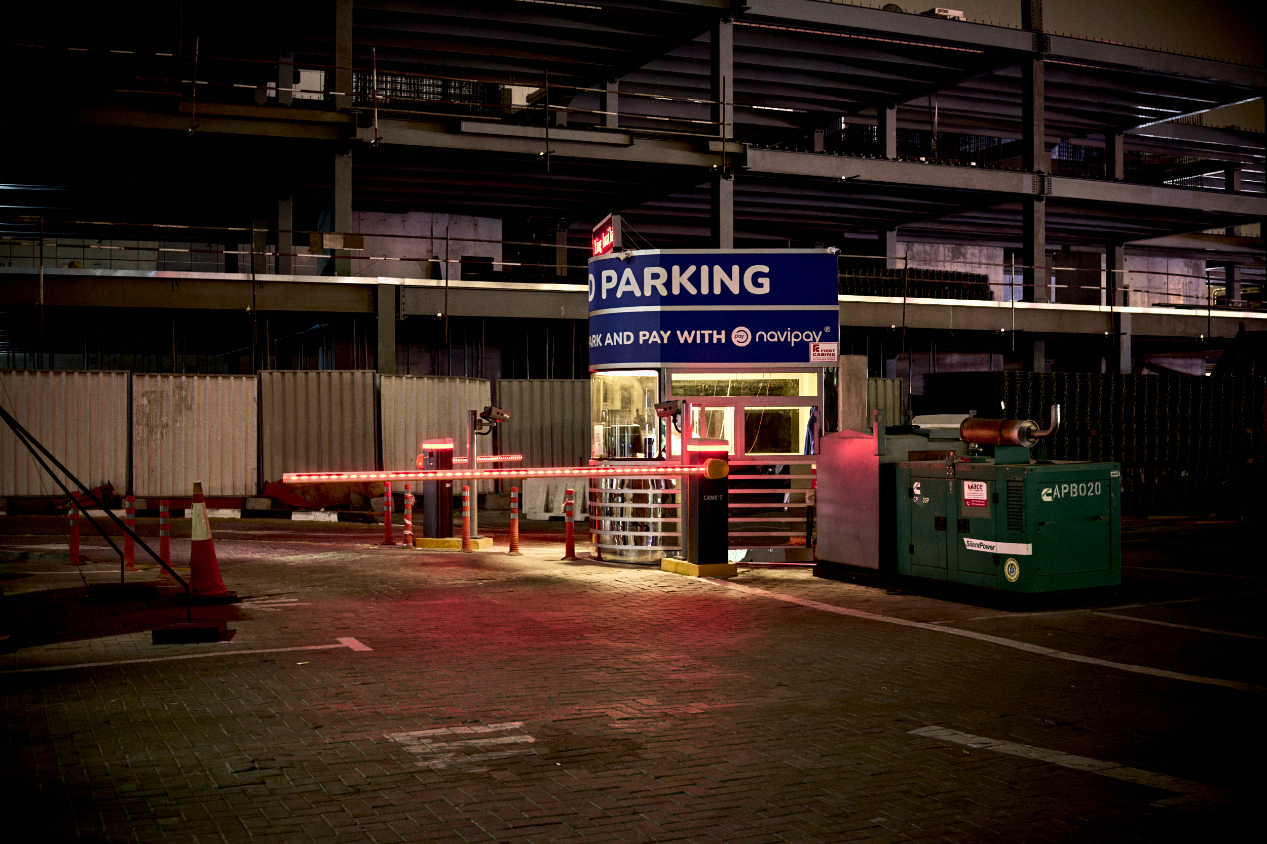 A parking lot entrance at night, with a lit barrier, payment booth, and PARKING sign overhead.