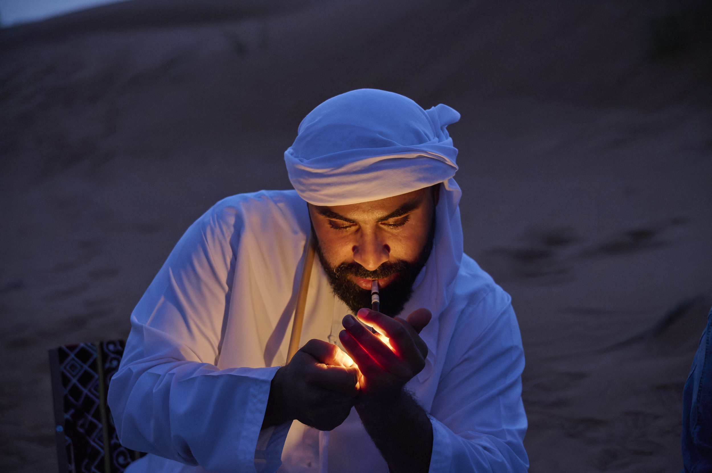 A man in traditional clothing lights a pipe, his face illuminated against a desert background at dusk.