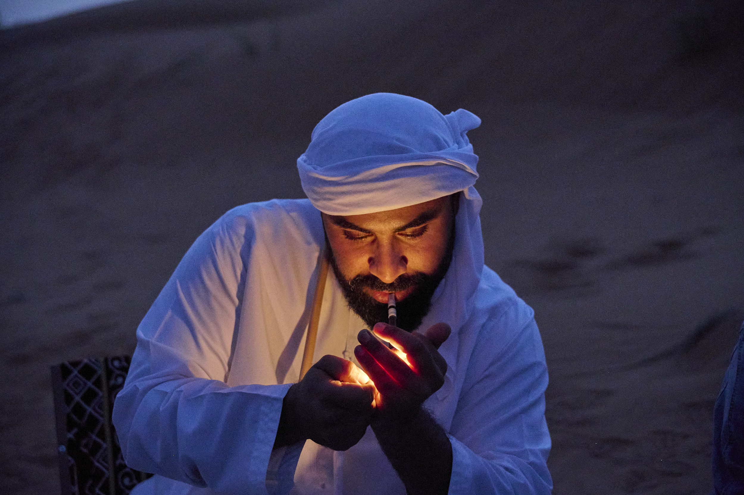 Man in traditional white attire lighting a pipe at dusk in a desert setting.