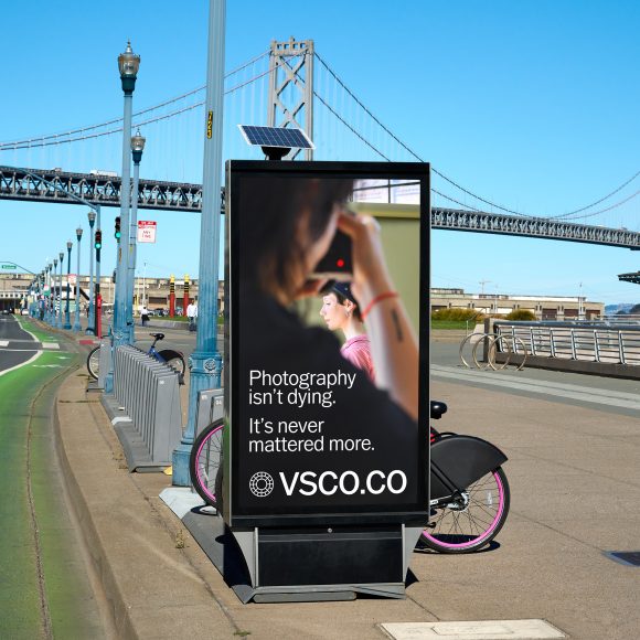 Digital billboard near a bridge shows a woman using a camera and text promoting VSCO photography.