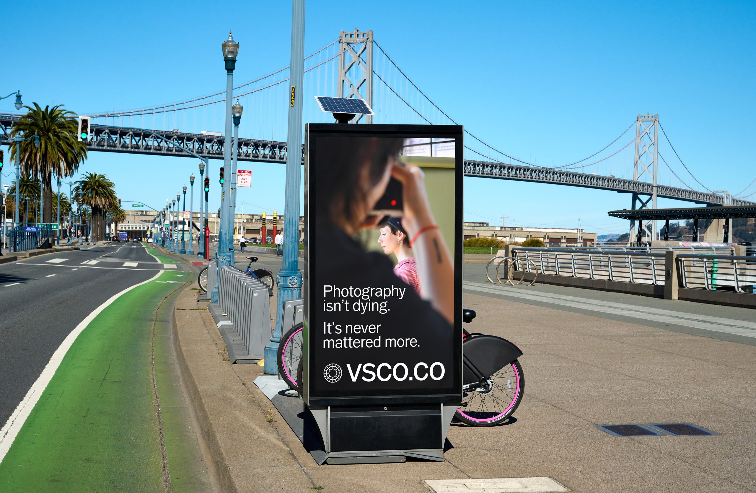 Digital billboard near a bridge shows a woman using a camera and text promoting VSCO photography.
