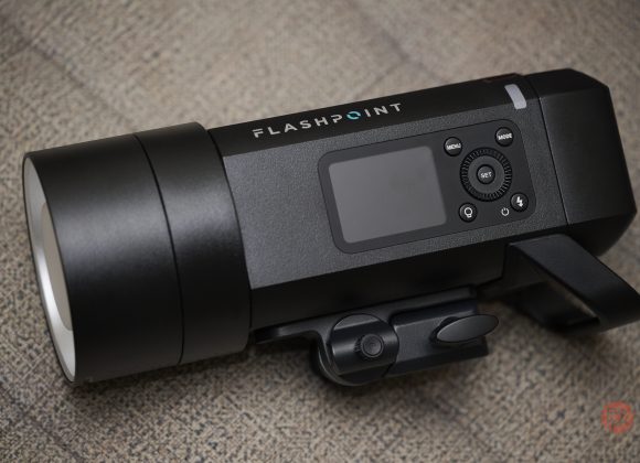 Flashpoint XPLOR 400 Pro II Strobe - An Unmissable Upgrade Black Flashpoint camera flash unit with control buttons and display, placed on a textured beige surface.