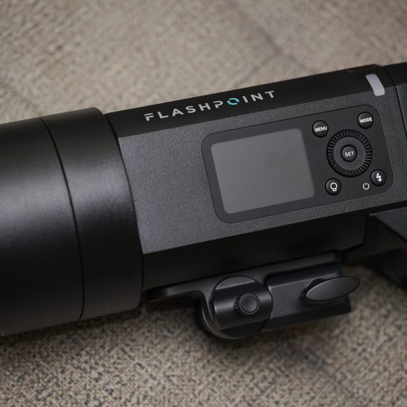 Black Flashpoint camera flash unit with control buttons and display, placed on a textured beige surface.