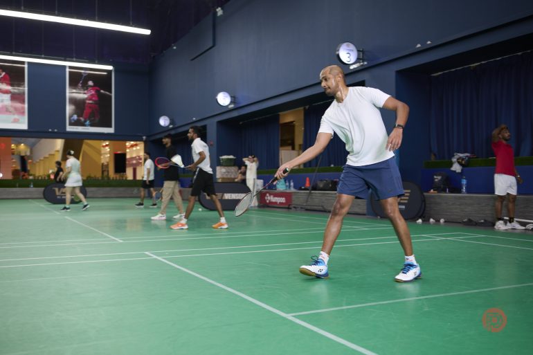 Four men play badminton indoors on a green court, with one player ready to hit the shuttlecock.