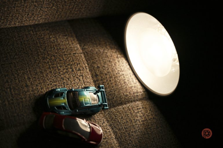 Two toy cars on a brown fabric surface beside a lit lamp, casting shadows.
