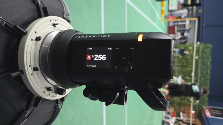 A Flashpoint camera flash unit displays settings on its screen at an indoor sports venue with a green floor.