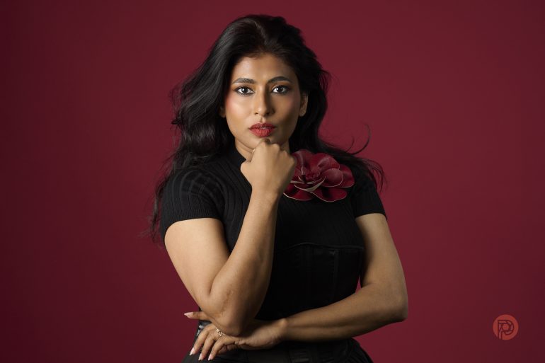 Woman with long dark hair poses confidently against a maroon background, wearing a black top with a flower accessory.