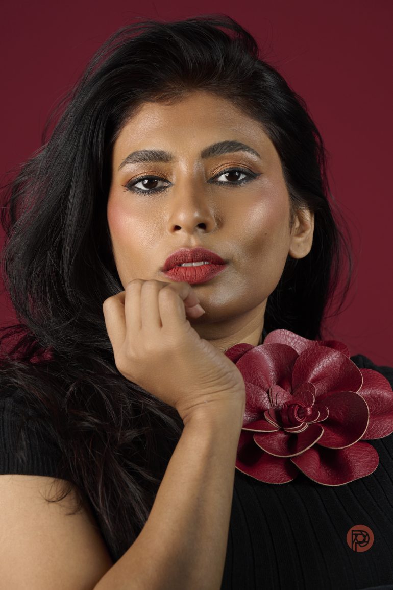 Woman with long dark hair and red lipstick posing against a red background, wearing a flower-shaped accessory.