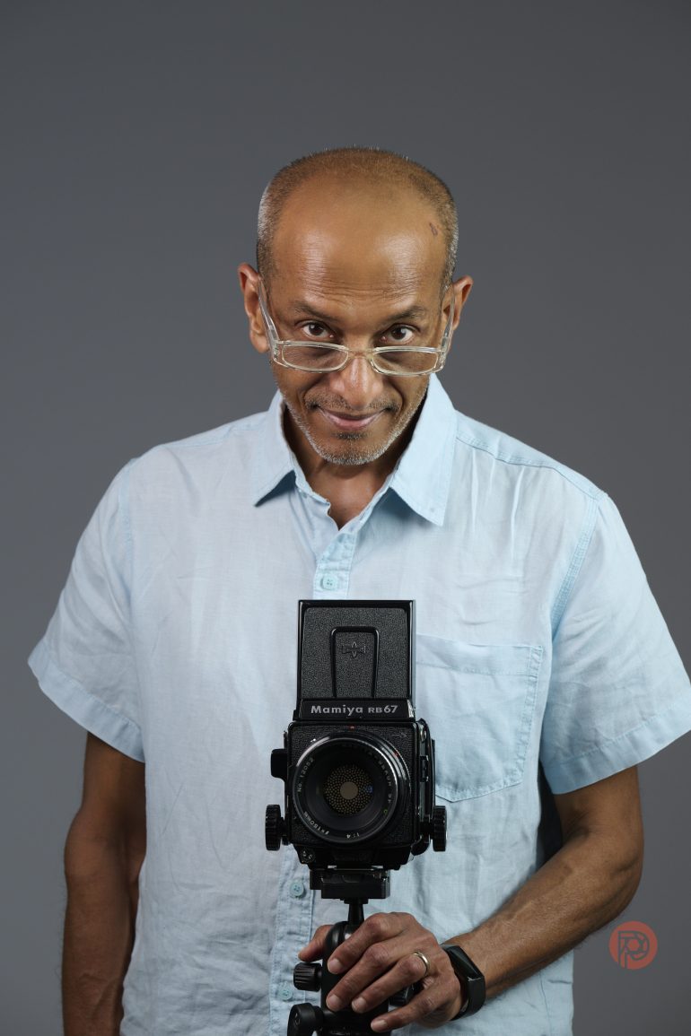 Bald man in glasses and light blue shirt holding a vintage camera, looking intently at the viewer.