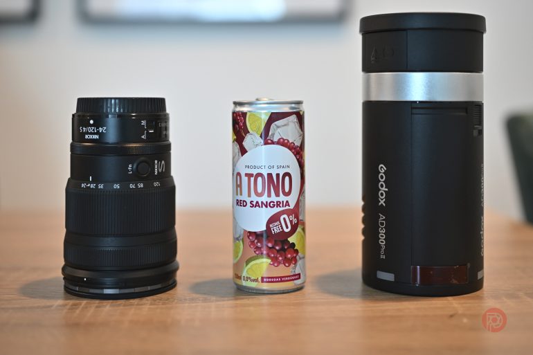 A camera lens, a can of Atomo Red Sangria, and a Godox flash unit on a wooden table.