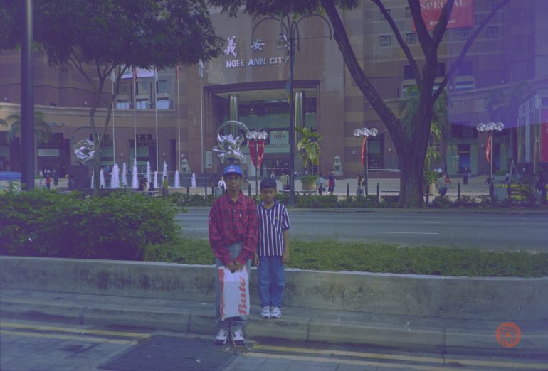 Two boys stand on a sidewalk in front of Ngee Ann City mall, with trees and fountains in the background.
