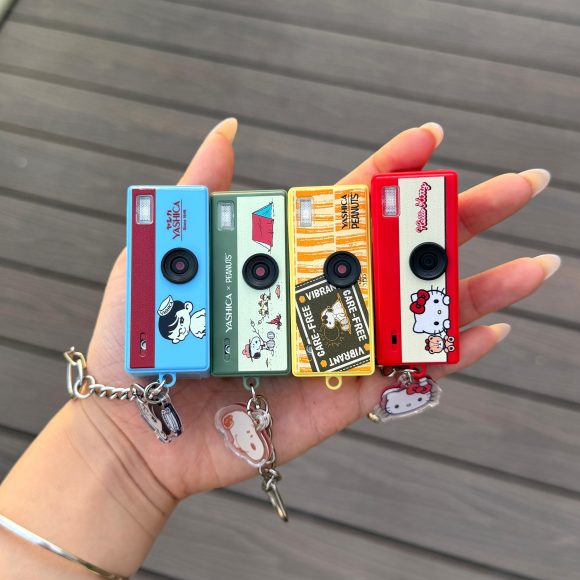 A hand holds four colorful toy cameras with cartoon designs, each on a keychain, over a wooden surface.
