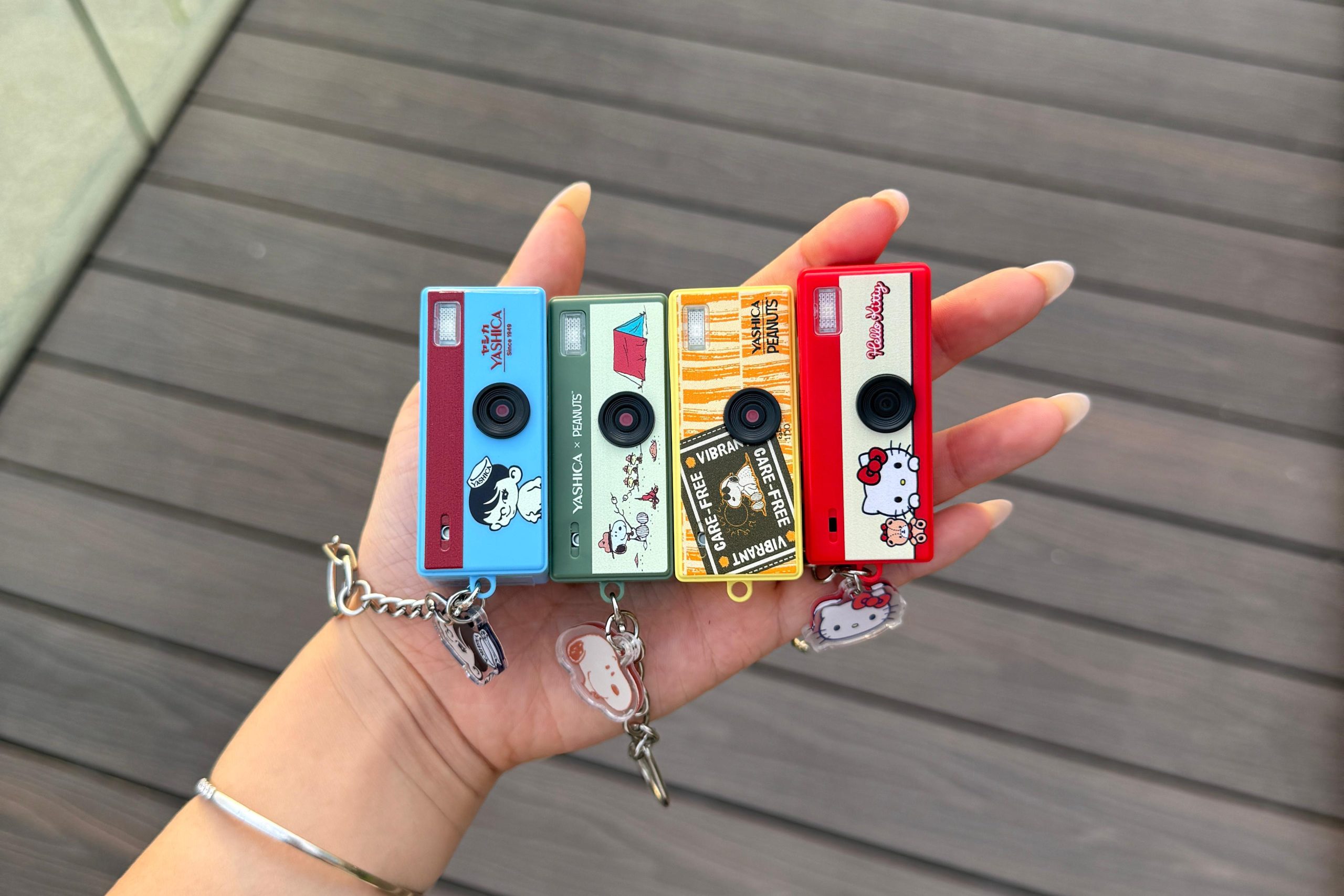 A hand holds four colorful toy cameras with cartoon designs, each on a keychain, over a wooden surface.