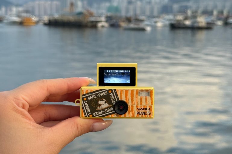 A hand holds a yellow Yashica camera with a small screen, near a harbor with boats and a city in the background.