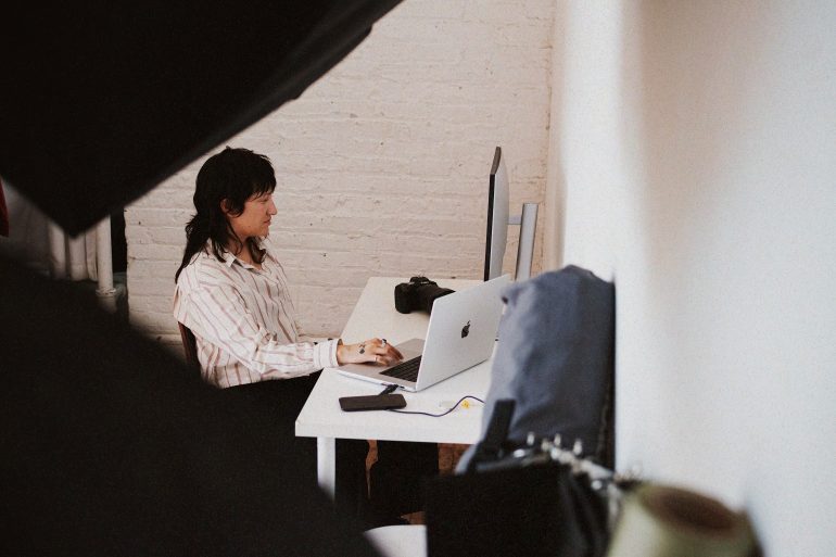 Person in striped shirt working at a desk with a computer and camera, seen through a partially obscured view.