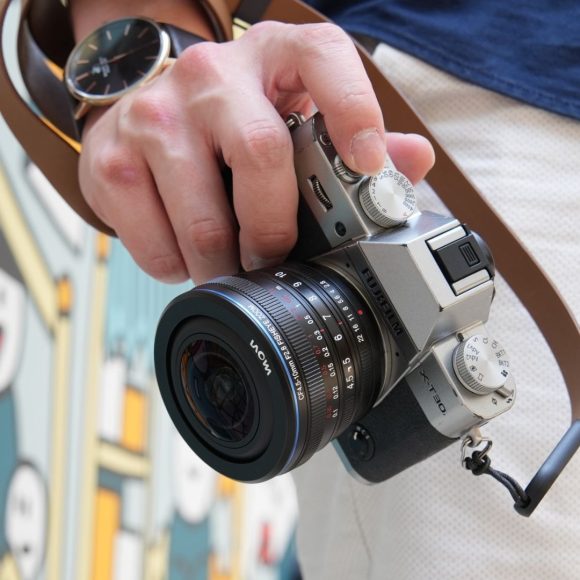 A person holding a silver camera with a brown strap, standing near a colorful mural.