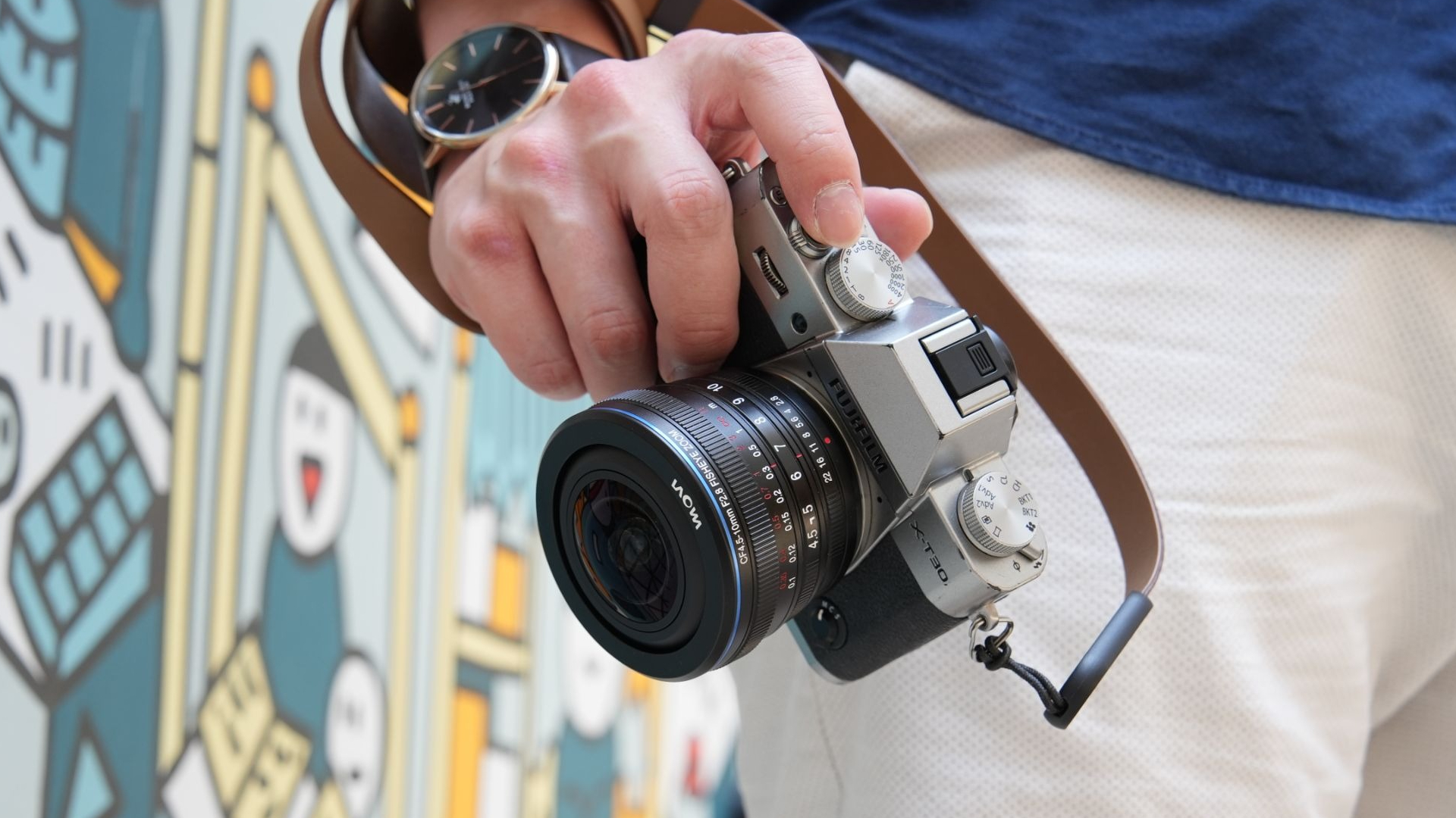 Laowa Just Made Fisheye Photography More Versatile Than Ever Before A person holding a silver camera with a brown strap, standing near a colorful mural.