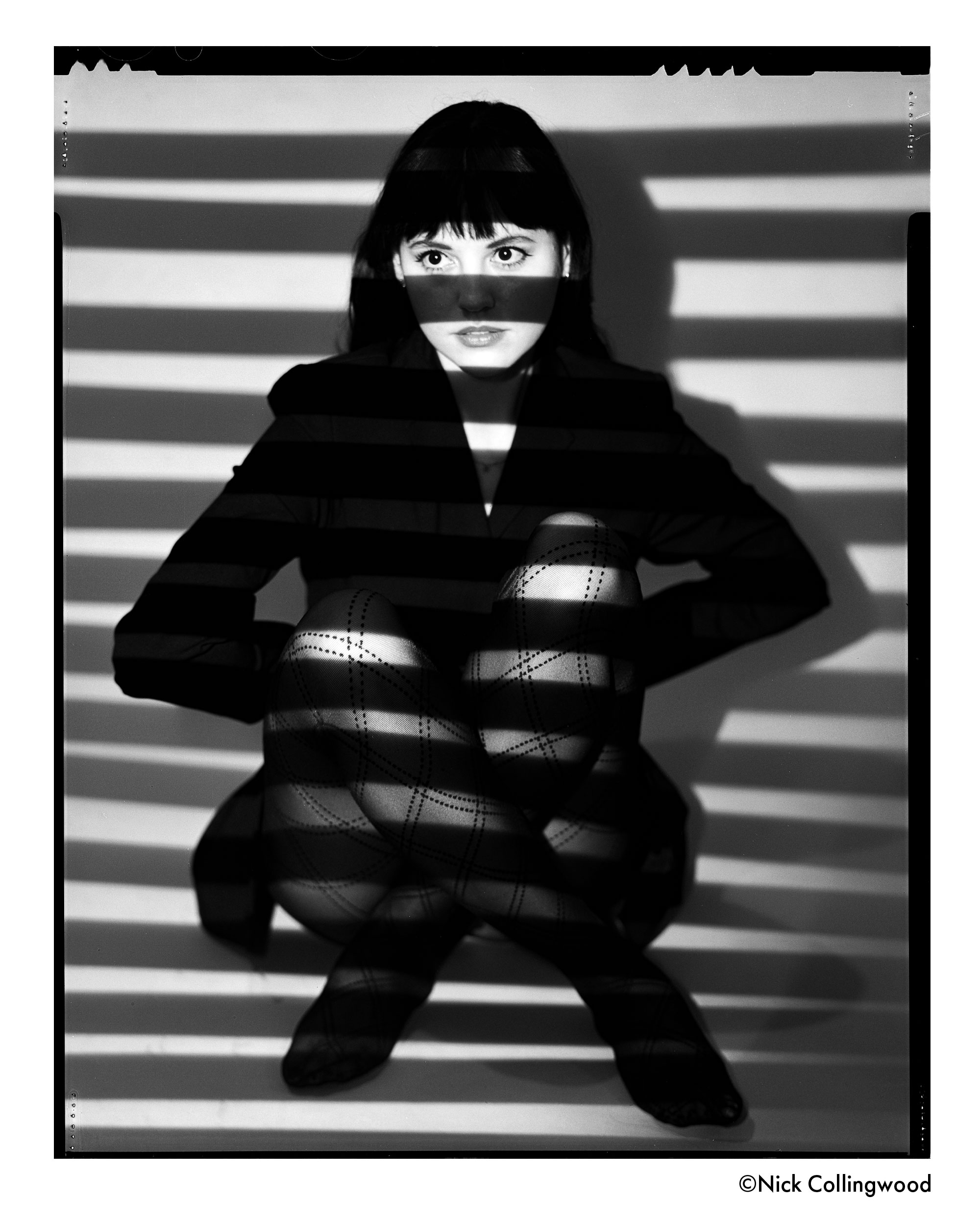 Woman seated with striped light and shadows across her face and body, wearing a dark blazer and patterned tights.