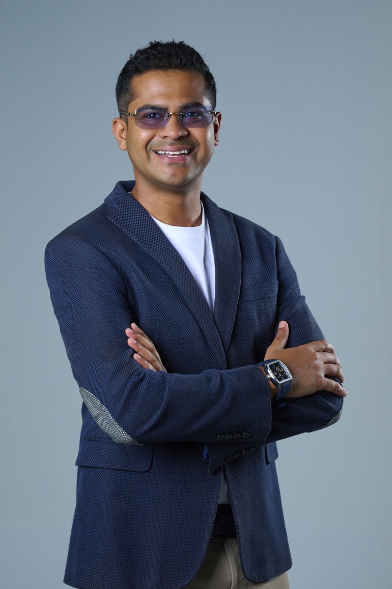 Smiling man in glasses and a blazer stands with arms crossed against a plain gray background.