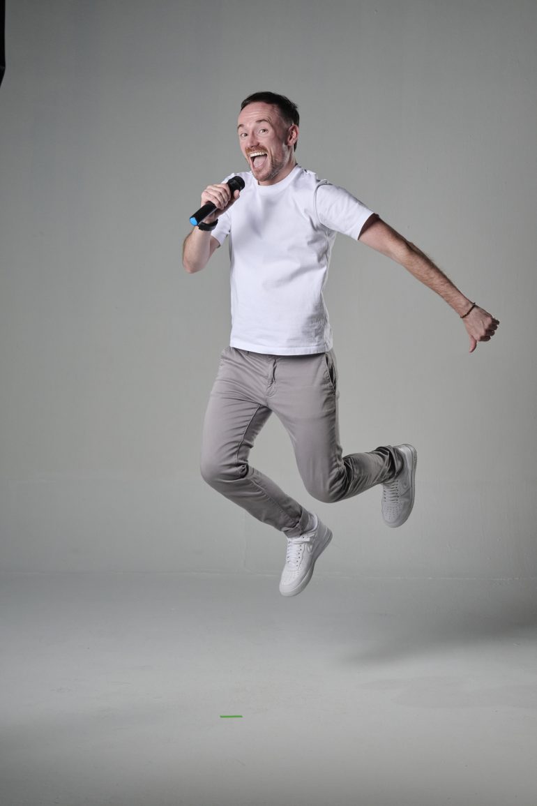 Man in white t-shirt and gray pants jumping while singing into a microphone against a plain background.