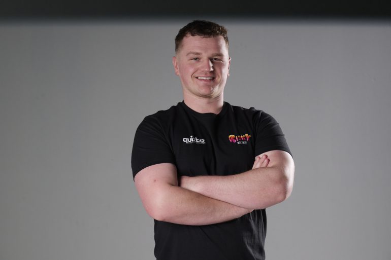 Man in a black t-shirt with arms crossed, smiling, standing against a plain light gray background.