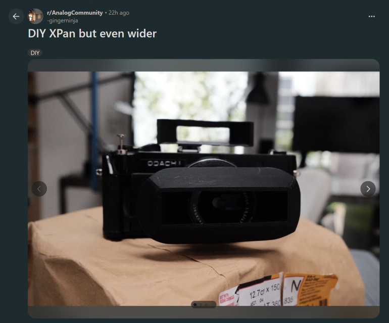 A black DIY panoramic film camera with a wide lens adapter sits on a brown package indoors.