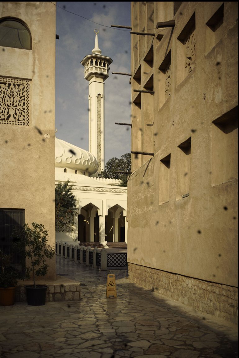 A narrow alley with beige buildings leads to a mosque with a white dome and tall minaret in the background.
