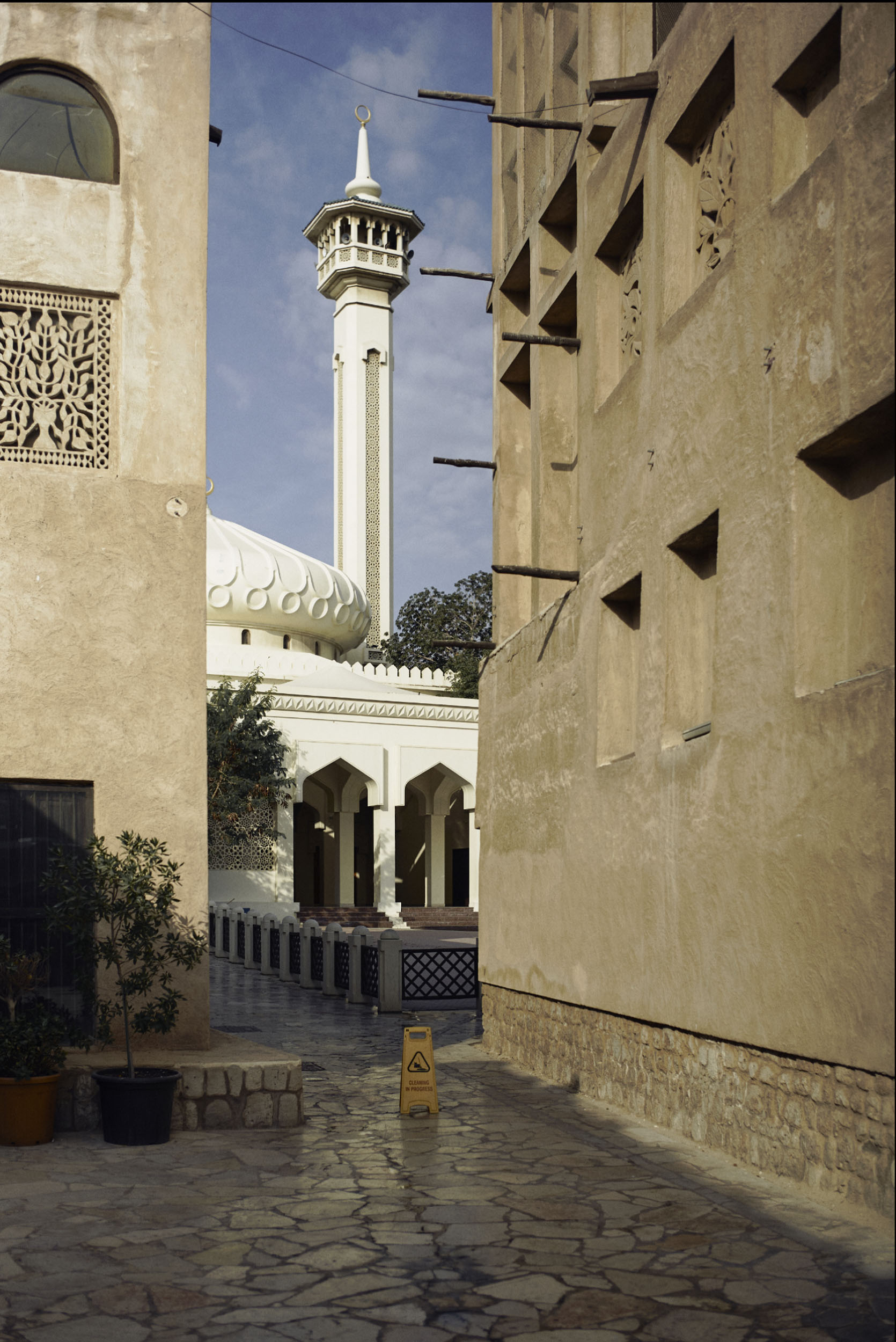 A mosque with a white dome and minaret seen through a narrow alley with beige buildings and a wet floor sign.