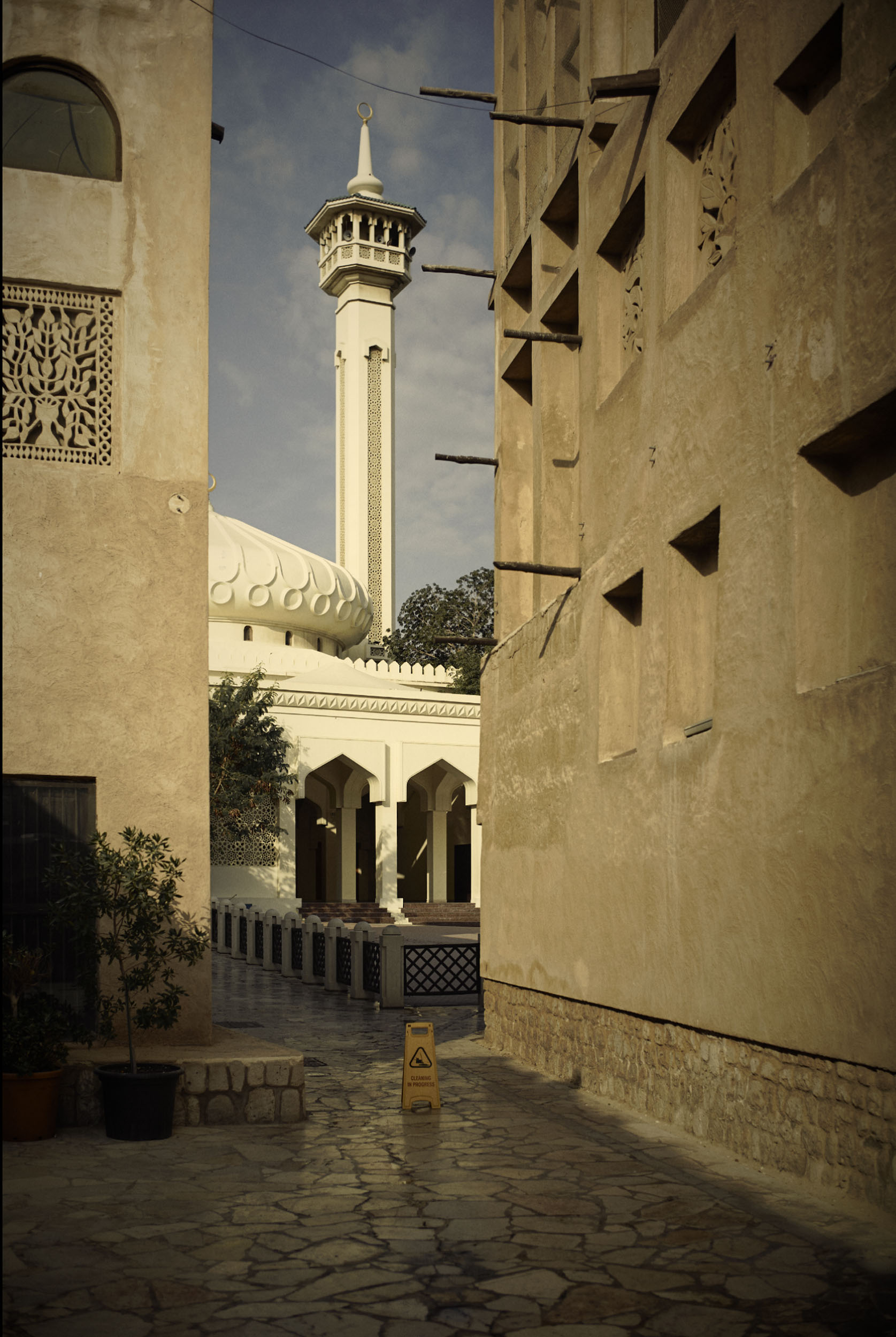 A narrow stone alley leads to a mosque with a tall minaret and dome, under a partly cloudy sky.