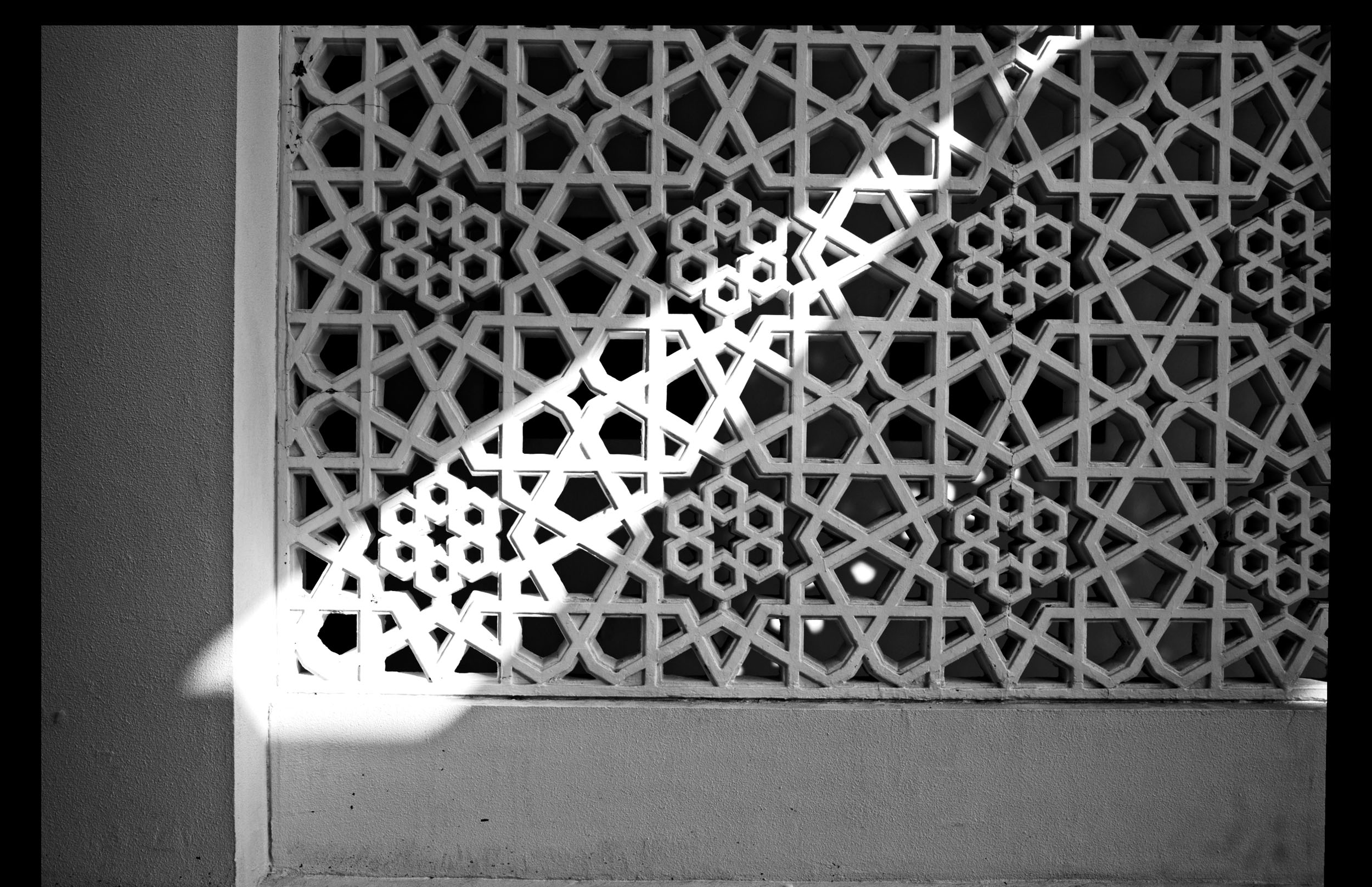 A patterned white lattice screen with a diagonal beam of sunlight casting shadows across it.