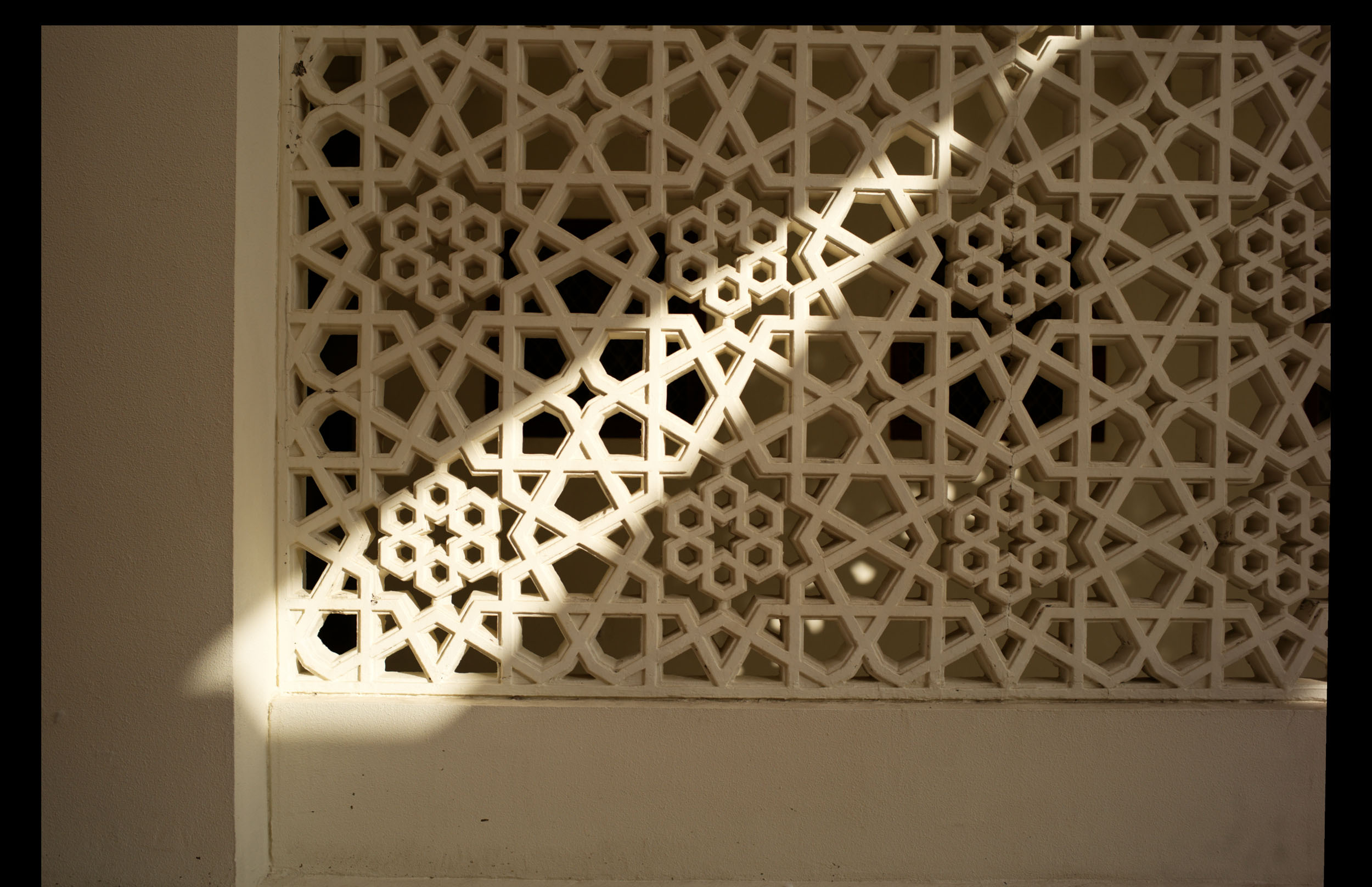 Sunlight casts a diagonal shadow on an ornate white lattice wall with geometric patterns.