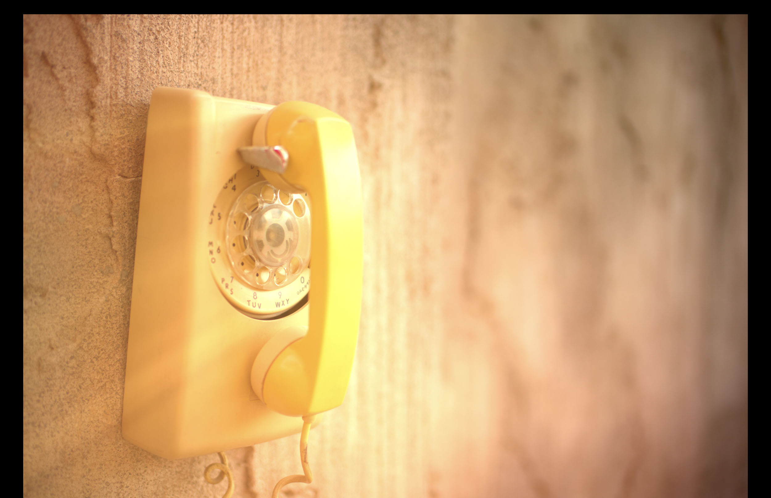 A yellow rotary dial telephone is mounted on a textured beige wall, lit by warm sunlight.