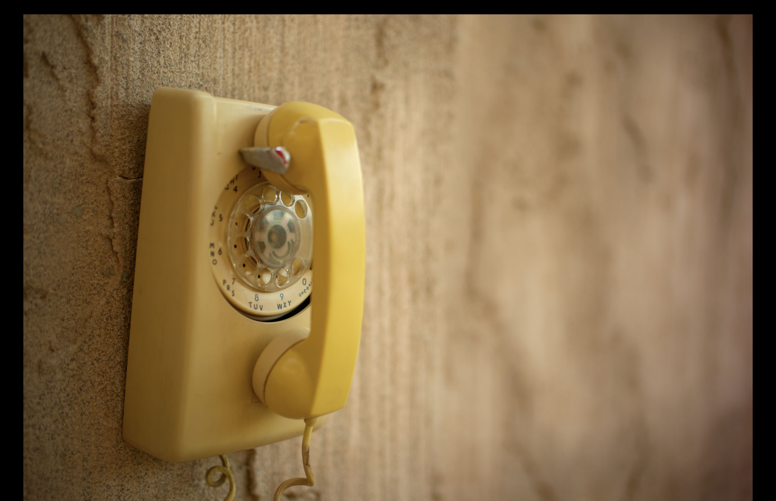 Yellow rotary dial wall phone hanging on a textured beige wall.
