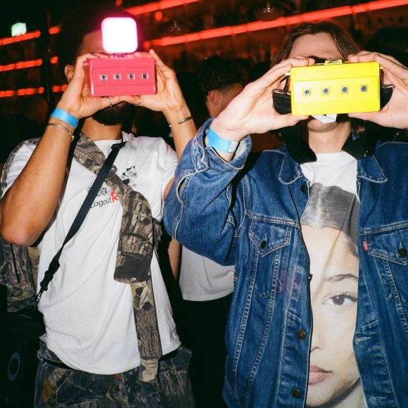 Two people at a party hold up colorful toy cameras, with bright lights and a crowd in the background.