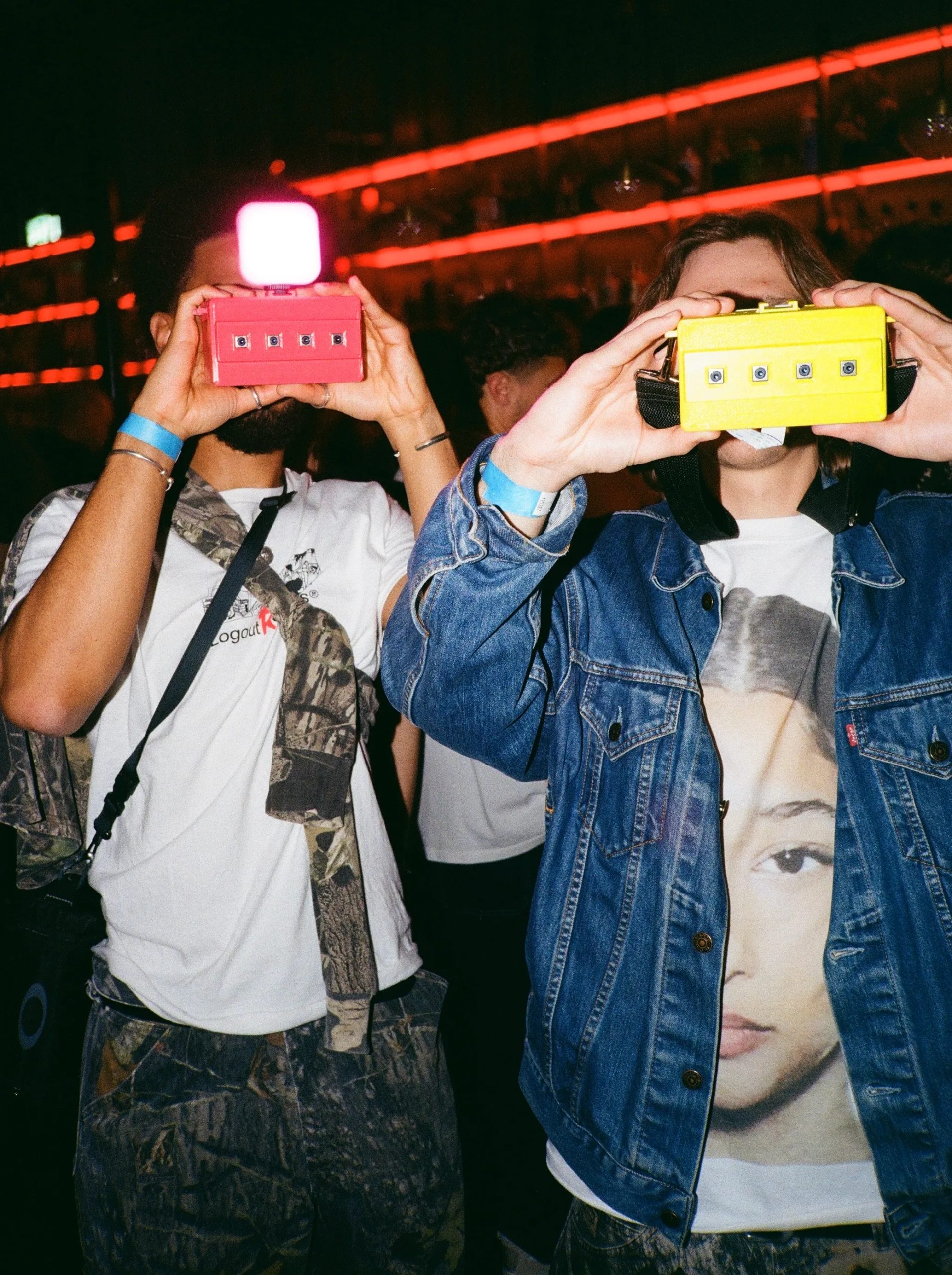Two people at a party hold up colorful toy cameras, with bright lights and a crowd in the background.
