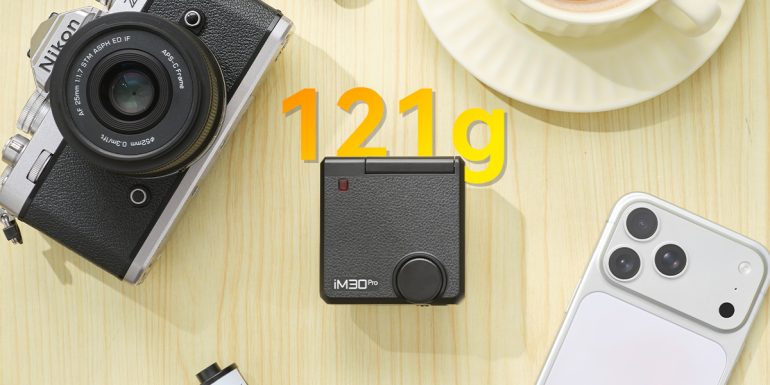 Flat lay of a camera, a phone, a coffee cup, and a compact black device labeled 121g on a wooden surface.