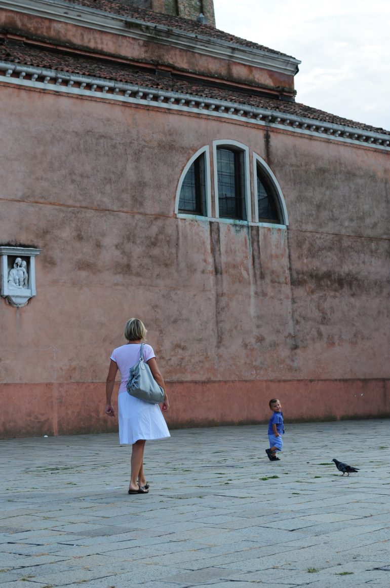 A woman and a child walk on a stone plaza near a large, old building with arched windows.