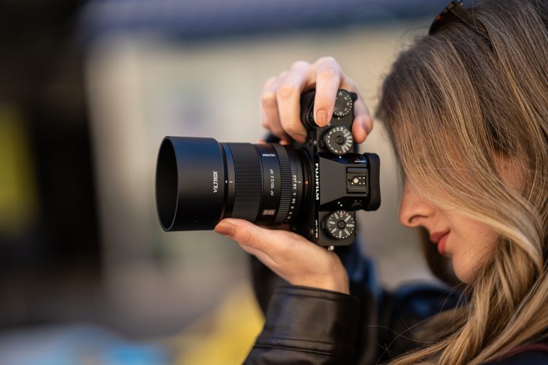 A woman with blonde hair holds a camera up to her eye, preparing to take a photo outdoors.