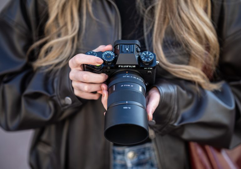 Person in a brown leather jacket holding a Fujifilm X-T5 camera with both hands.