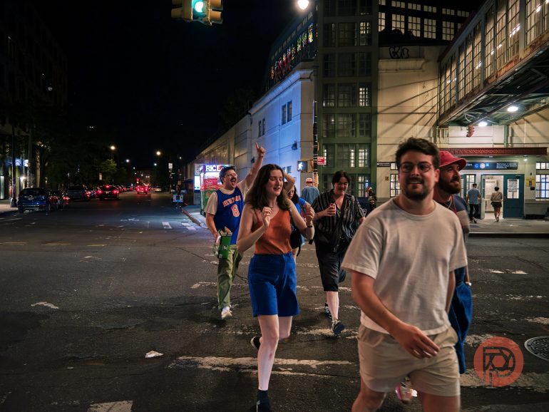 A group of young adults happily crossing a city street at night under bright lights.